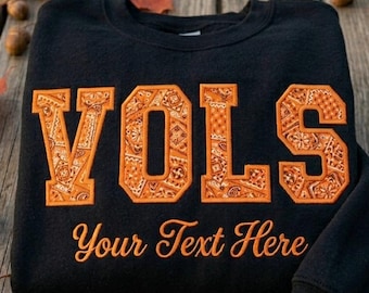 Könnte beinhalten: Ein schwarzes Sweatshirt mit Rundhalsausschnitt und dem Wort VOLS in großen Blockbuchstaben, die mit einem orangefarbenen Bandanamuster gefüllt sind. Darunter ist der Schriftzug Your Text Here in orangefarbener Schreibschrift gestickt.