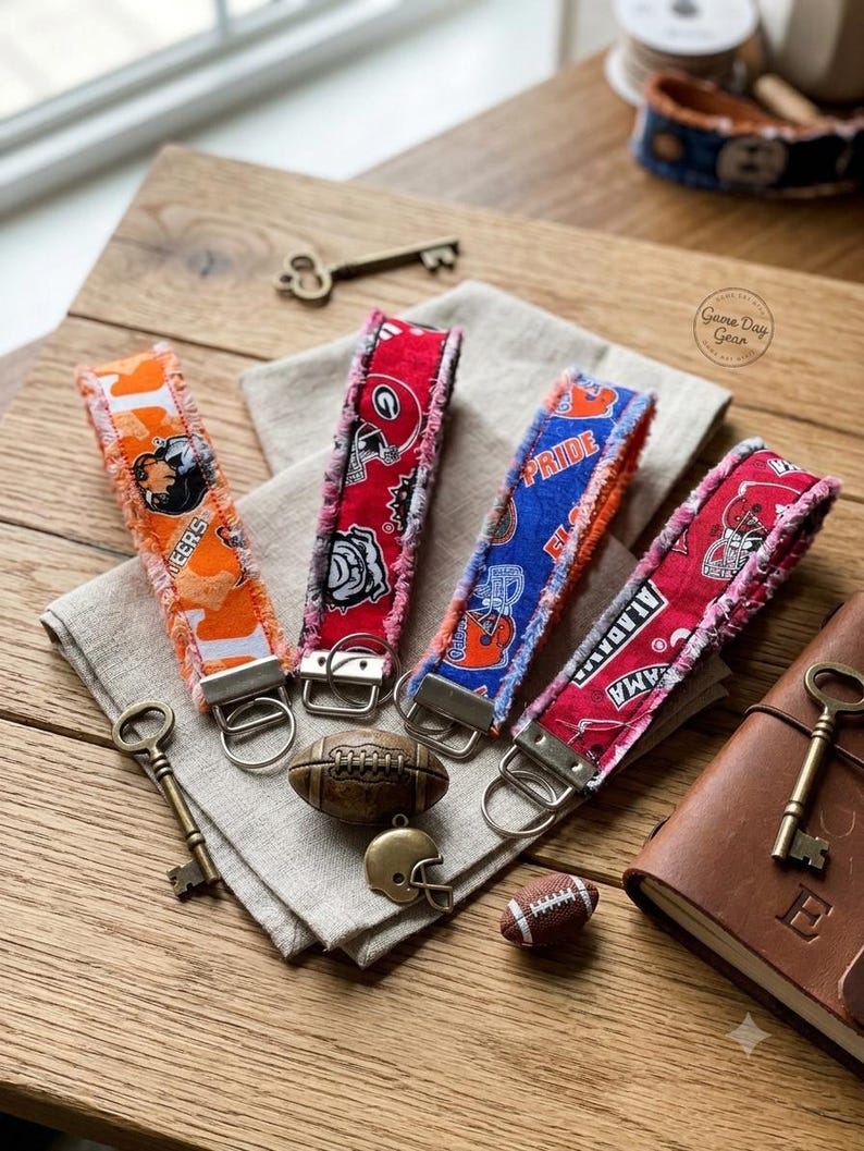 May include: Four fabric wristlet keychains with frayed edges, each featuring distinct college sports team patterns in orange, red, and blue. The wristlets include silver-toned metal hardware with attached split rings for keys.