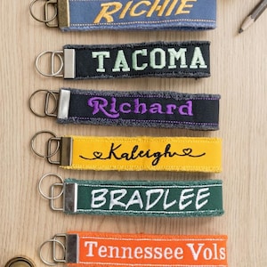 May include: Six fabric keychains with frayed edges and metal rings. The blue one says RICHIE, black says TACOMA, black says Richard, yellow says Kaleigh with hearts, green says BRADLEE, and orange says Tennessee Vols in white embroidery.