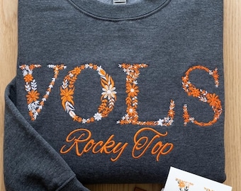 Peut inclure: Un sweat-shirt à col rond gris anthracite foncé avec le mot VOLS brodé en motifs floraux orange et blancs, et le texte Rocky Top brodé en dessous en écriture orange. Inclut une étiquette gland marron et une étiquette produit blanche.