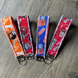 May include: Four fabric wristlet keychains with frayed edges and metal hardware. The first is orange with a mascot, the second is red with a logo, the third is blue with the text PRIDE and a helmet, and the fourth is red with the text ALABAMA.