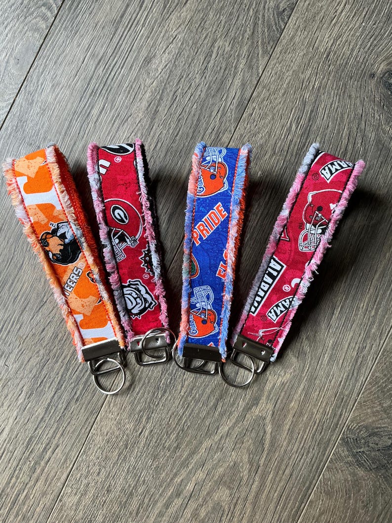 May include: Four fabric wristlet keychains with frayed edges and metal hardware. The first is orange with a mascot, the second is red with a logo, the third is blue with the text PRIDE and a helmet, and the fourth is red with the text ALABAMA.