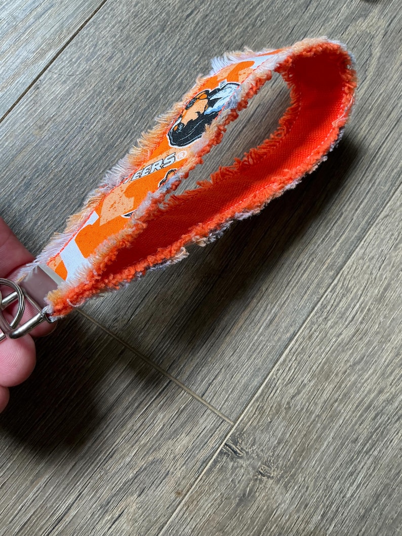 May include: A fabric wristlet keychain featuring a bright orange and white pattern with frayed edges. The strap displays a graphic of a black and white animal head and the partial text EERS in orange block letters, attached to a silver metal ring.