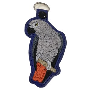 May include: A blue leather keychain with an embroidered gray parrot perched on a branch. The parrot has a yellow eye and a red foot.
