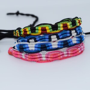 Puede incluir: Un conjunto de cuatro pulseras de amistad de colores. La pulsera superior es negra, verde, amarilla y roja. La segunda pulsera es azul, blanca y negra. La tercera pulsera es rosa y blanca. La pulsera inferior es rosa y blanca.