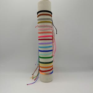 May include: A display of colorful friendship bracelets on a white stand. The bracelets are made of woven string and come in a variety of colors, including red, orange, yellow, green, blue, purple, pink, and black.