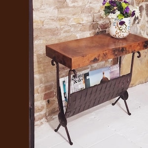 May include: A rustic, copper-topped side table with a wrought iron magazine rack. The table features a rectangular top and curved legs. A decorative vase with purple and white flowers sits on top. Magazines are visible in the rack.