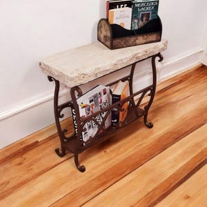 May include: A decorative console table with a natural stone top and a wrought iron base. The table features a lower shelf for magazines and books. The table is brown and beige and is placed on a hardwood floor.