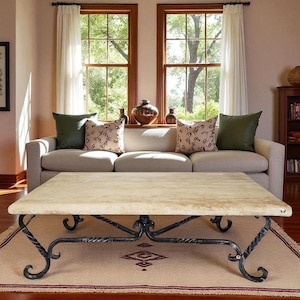 May include: A rectangular coffee table with a light-colored stone top and ornate black metal legs. The table sits on a patterned rug in front of a beige sofa with decorative pillows. Two windows with white curtains are in the background.