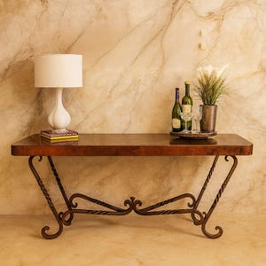 May include: A dark brown console table with wrought iron legs. A white lamp, books, wine bottles, glasses, and a potted plant are displayed on the table. The background is a beige marble wall.