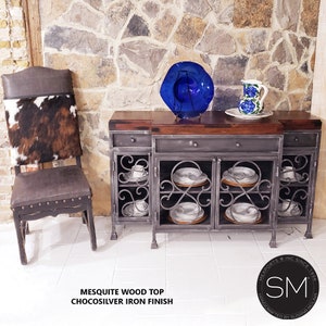 May include: A rustic wooden top cabinet with a chocolate silver iron finish. The cabinet has two doors with glass panels and a decorative iron design. The cabinet is sitting on a white tiled floor. The cabinet is decorated with a blue glass bowl and a white pitcher with blue flowers.