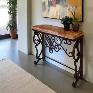 May include: A decorative console table with a marble top and ornate wrought iron base. The tabletop has a reddish-brown hue. The iron base features scrolled and geometric designs. The table is placed against a neutral wall, with a painting and potted plants.