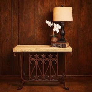 May include: A decorative side table with a light-colored stone top and ornate, wrought-iron base. A black lamp with a beige shade, a small vase with white flowers, and a vintage book are displayed on the table. The table is set against a dark wood-paneled wall.