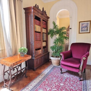 May include: A room featuring a large, ornate wooden bookcase filled with books. A burgundy armchair sits on a patterned rug, and a small table with a decorative base is in the foreground. A potted plant and a framed picture complete the scene.