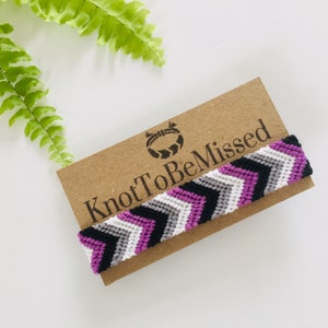 Op de afbeelding: Een handgemaakte vriendschapsarmband met een chevronpatroon in de kleuren van de aseksuele pride-vlag: zwart, grijs, wit en paars. De armband wordt gepresenteerd op een bruine kaart met de tekst "KnotToBeMissed".