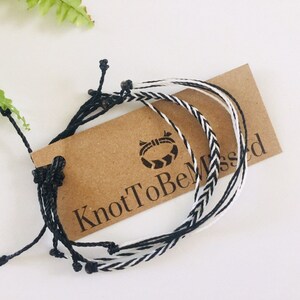 May include: Set of three black and white friendship bracelets with black beads. The bracelets are tied together and are on a brown card with the text "KnotToBeMissed".