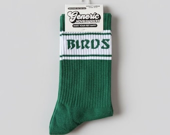 Peut inclure: Une paire de chaussettes montantes côtelées vertes avec une bande horizontale blanche près du haut, portant le mot BIRDS imprimé en police gothique vert foncé. L'étiquette en carton blanc affiche le texte Generic NOVELTY SOCKS MAKE YOUR FEET HAPPY.