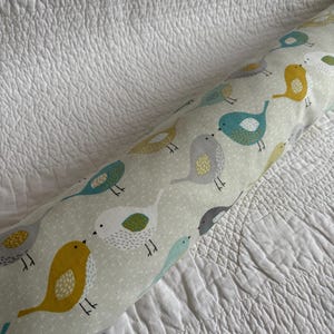 May include: A cylindrical bolster cushion with a repeating pattern of colourful birds in yellow, teal, grey, and white on a light green background. The birds are stylized with simple shapes and dots. The cushion is on a white, textured surface.