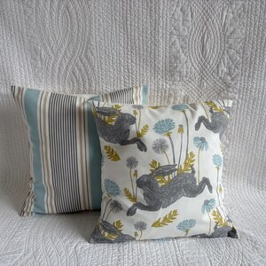 May include: Two square scatter cushions. One cushion features a vertical striped pattern in shades of light blue, white, and grey. The other cushion displays a white background with a repeating pattern of grey hares, yellow leaves, and blue flowers.