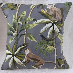 May include: A grey throw pillow with a tropical print featuring green palm trees and two monkeys. One monkey is brown and sitting on a branch, while the other monkey is grey and hanging from a branch.