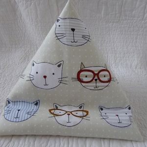 May include: A triangular beige pillow with a white polka dot pattern featuring five cartoon cats with different expressions. The cats have black eyes, white fur, and various details like glasses, stripes, and a red nose.
