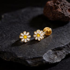 Puede incluir: Un par de pendientes de botón dorados con forma de margarita, con pétalos blancos y centros amarillos. Los pendientes se exhiben sobre una superficie oscura y texturizada, con un fondo oscuro. Una pequeña roca marrón oscura en el fondo.