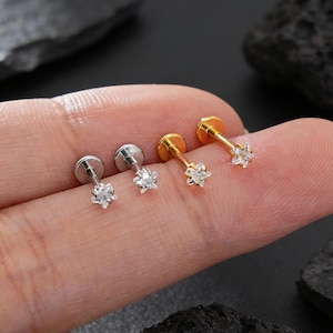 Puede incluir: Cuatro pendientes pequeños de botón. Dos son de color plateado y dos dorados. Cada pendiente presenta una gema transparente en forma de estrella. Los pendientes se muestran en un dedo sobre un fondo oscuro con rocas.