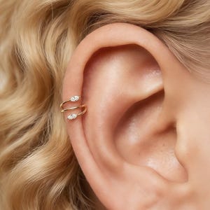 Pode incluir: Um ear cuff de tom dourado com um design em espiral de banda dupla. As extremidades da peça são acentuadas com duas pequenas pedras transparentes de corte marquise. O acessório está posicionado na cartilagem superior da orelha.