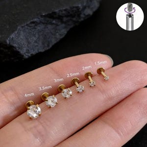 Op de afbeelding: Een verzameling goudkleurige labret studs met heldere, ronde edelstenen. Maten variëren van 1,5 mm tot 4 mm. De afbeelding toont ook een close-up van het interne schroefdraadmechanisme van de labret stud.