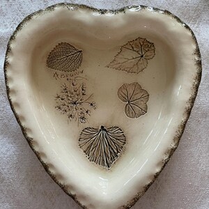 May include: Heart-shaped ceramic dish with a cream-colored interior and a textured, brown rim. The dish features leaf imprints in shades of brown and the text "Brown". A decorative piece for serving or display.