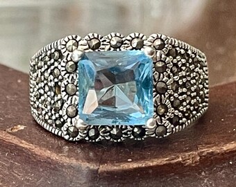 May include: A square-cut, light blue gemstone ring. The ring has a silver band with a detailed pattern of small, dark stones. The gemstone is the focal point, set in a decorative silver setting.
