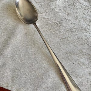 May include: A silver spoon with a long handle and an oval bowl. The spoon is made of a shiny, reflective metal and is resting on a textured, light-colored surface. The spoon is likely used for serving or eating.