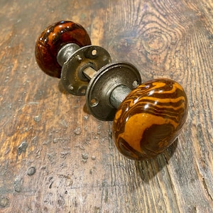 May include: A pair of round door knobs featuring a marbled pattern of dark brown and amber swirls. The knobs are connected by a central metal spindle and include two circular metal mounting plates with screw holes, finished in a weathered gray tone.
