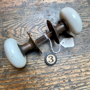 May include: A vintage door knob set featuring two smooth, white porcelain knobs connected by a central metal spindle and two circular metal plates. A small white paper tag with the handwritten text A3 is attached, and a black circular token with the number 3.