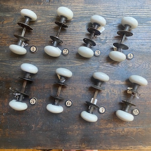 May include: Eight white porcelain door knobs with dark metal hardware and square spindles. Each set includes two knobs connected by a metal rod and circular mounting plates. Each unit is labeled with a small circular tag numbered one through eight.