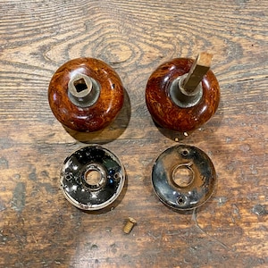 May include: Two round, brown marbled door knobs with metallic centers, one featuring a square spindle. Two circular black metal backplates with screw holes and a single small metal screw are positioned below the knobs on a wooden surface.