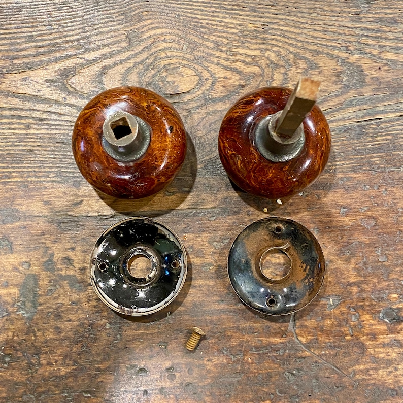 May include: Two round, brown marbled door knobs with metallic centers, one featuring a square spindle. Two circular black metal backplates with screw holes and a single small metal screw are positioned below the knobs on a wooden surface.