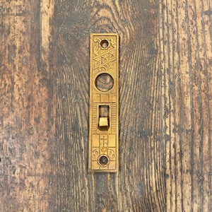 May include: A rectangular, antique brass door plate with ornate, engraved designs. The plate features a keyhole, a switch, and decorative patterns. The brass has a warm, aged patina, suggesting its vintage origin. The plate is mounted on a wooden surface.