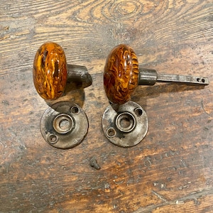 May include: A pair of round door knobs featuring a marbled amber and brown swirl pattern with a glossy finish. Each knob is attached to a dark metallic spindle and is accompanied by a circular metal mounting plate with three screw holes.