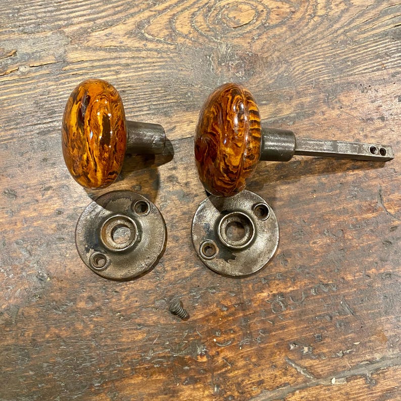 May include: A pair of round door knobs featuring a marbled amber and brown swirl pattern with a glossy finish. Each knob is attached to a dark metallic spindle and is accompanied by a circular metal mounting plate with three screw holes.