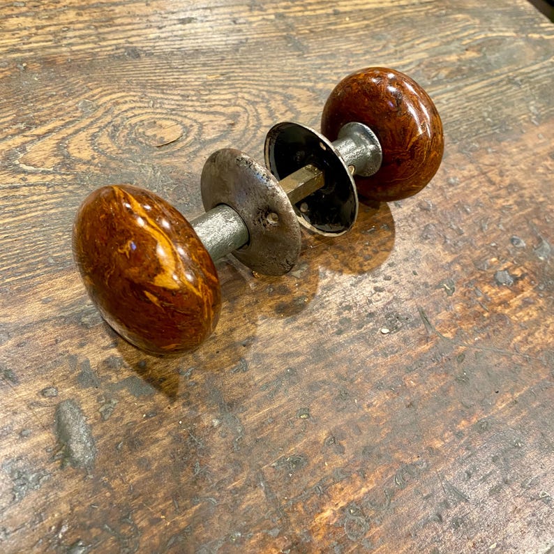 May include: A vintage door knob set featuring two round, polished brown and amber marbled knobs. The knobs are connected by a central metal spindle and include two circular metal backplates with visible screw holes and a weathered silver finish.