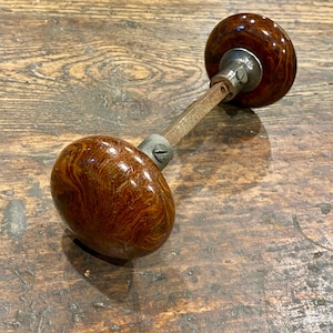 May include: A door knob set featuring two round, polished brown marbled knobs connected by a square metal spindle. Each knob has a metallic base with a visible screw, showcasing a swirling wood-grain pattern in shades of dark and light brown.