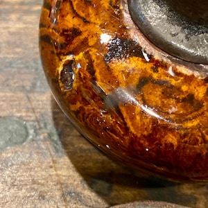 May include: A round, polished door knob featuring a glossy, marbled finish with swirling patterns of dark brown, amber, and golden yellow hues. The surface has a reflective, smooth texture with a dark metallic base visible at the top edge.