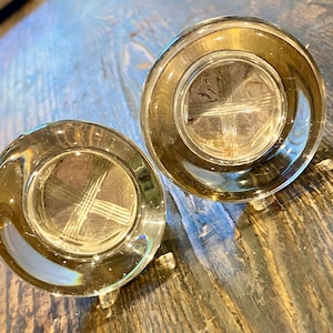 May include: Two clear glass candle holders with a gold rim and a cross design on the bottom. The candle holders are on a wooden surface.