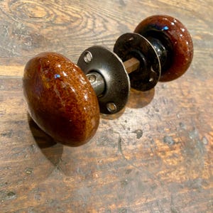 May include: A door knob set featuring two round, glossy, brown ceramic knobs with a mottled finish. The knobs are connected by a central metal spindle and include two circular, dark metal mounting plates with visible screw holes for installation.