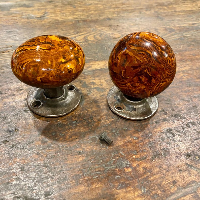 May include: Two round door knobs with a glossy, marbled brown and amber finish. Each knob is attached to a circular, dark grey metal base plate with two screw holes. A single small metal screw is positioned between the two door knobs.