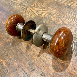 May include: A pair of round, brown, marbled-pattern door knobs connected by a central metal spindle. The knobs feature a glossy finish and are attached to circular metal backplates with a weathered, grey, metallic appearance and visible screws.