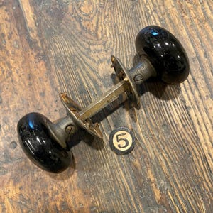 Puede incluir: Un juego de pomos de puerta vintage con dos pomos esféricos de color negro brillante conectados por un eje metálico central y dos placas de montaje circulares desgastadas. Una pequeña ficha redonda en blanco y negro con el número 5 está al lado.