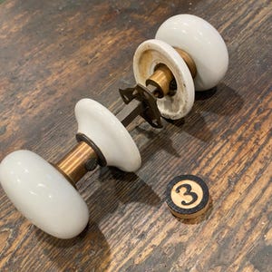 May include: A door knob set featuring two smooth, white, rounded porcelain knobs connected by a central brass spindle and a metal mounting plate. A small, circular, light-colored wooden disc with the black number 3 printed on it sits nearby.