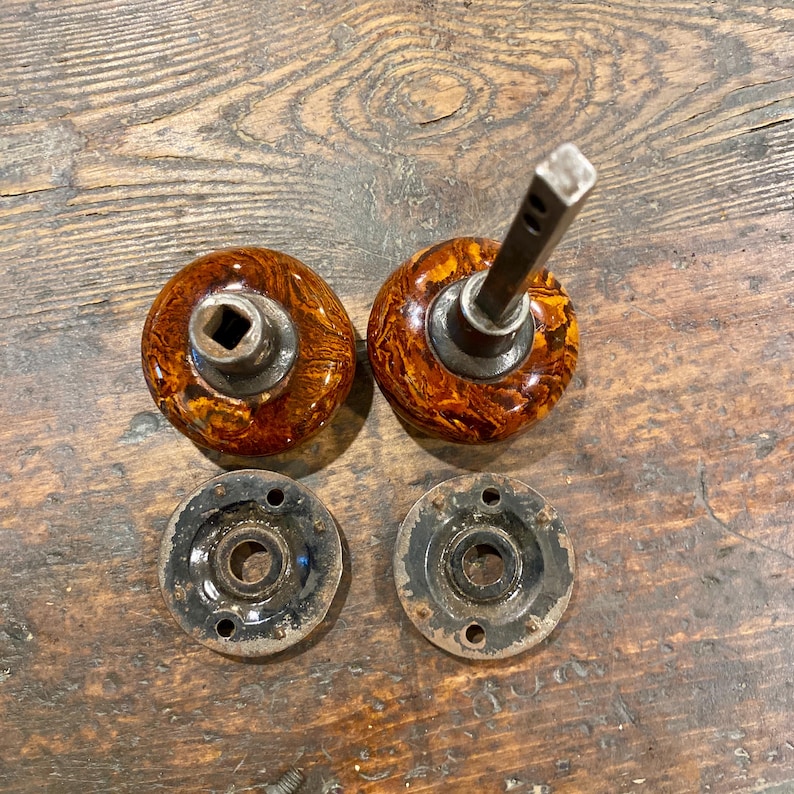 May include: Two round door knobs with a marbled brown and amber finish, featuring dark metal centers and a square spindle. Included are two circular black metal mounting plates with three screw holes each, designed for door hardware installation.